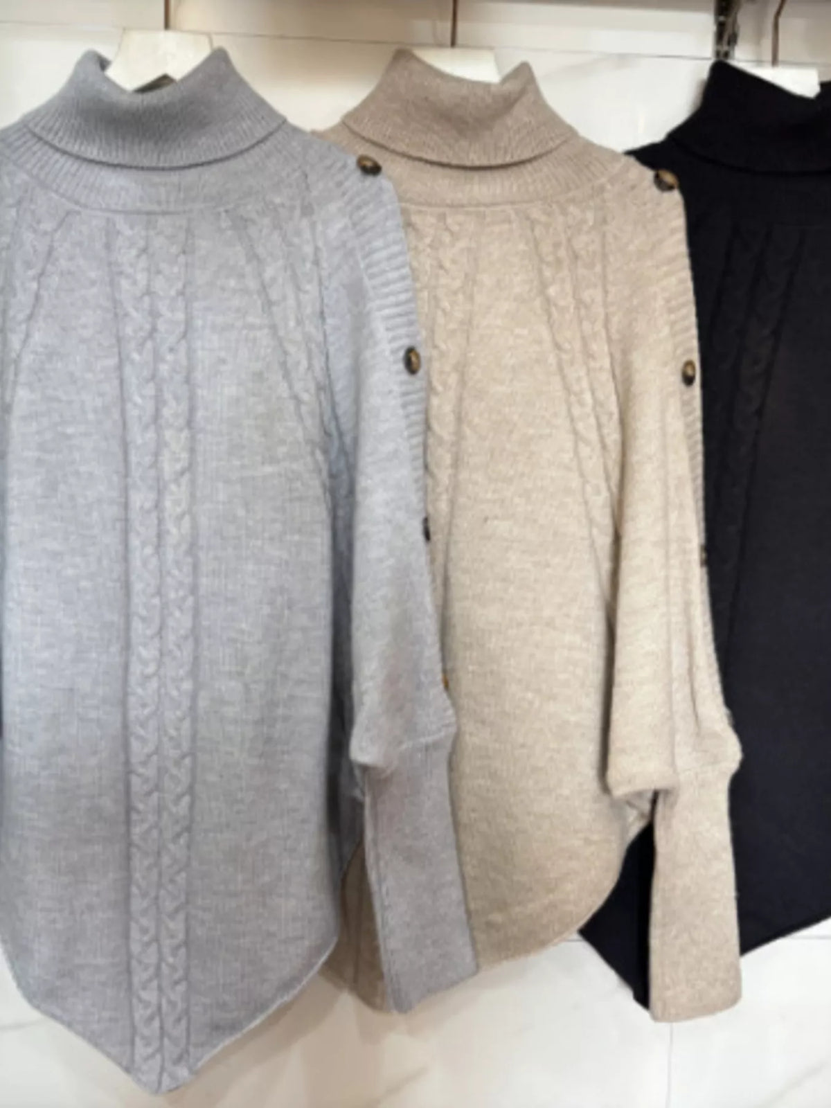 Light gray, warm beige/oatmeal, black cable-knit turtleneck cape sweater. Flat-lay/garment grouping of three cape sweaters. Styling accessories are not included.