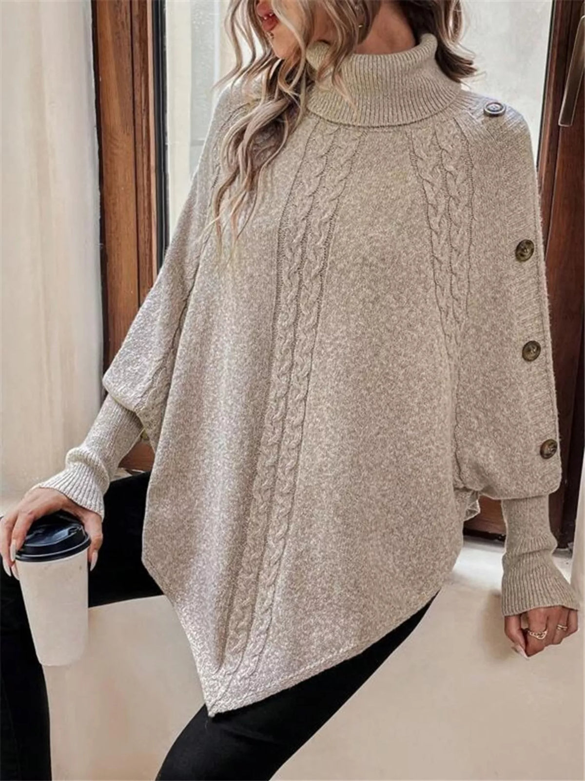 Warm beige/oatmeal cable-knit turtleneck cape sweater, front view with high neck and shoulder buttons. Styling accessories are not included.