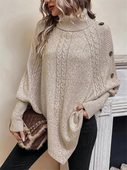 Warm beige/oatmeal cable-knit turtleneck cape sweater. Front three-quarter view with purse; model in black pants. Styling accessories are not included.