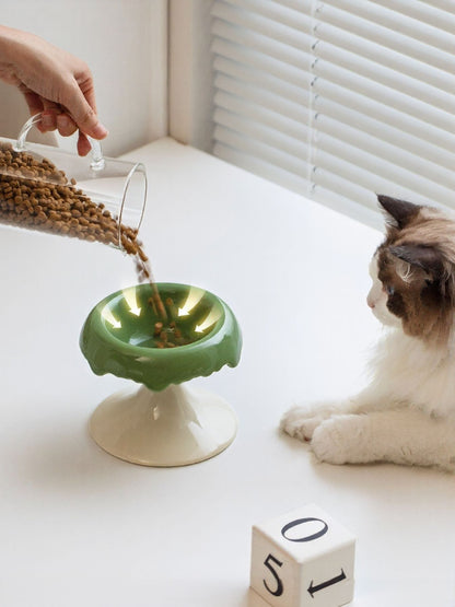 Green-and-white elevated bowl shown as food is poured, with a cat beside it.