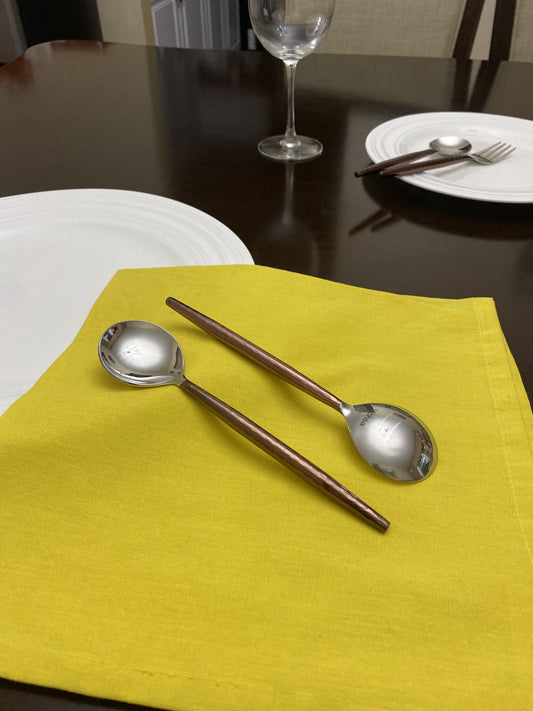 Two Vibhsa stainless steel soup spoons with hammered brown handles on a yellow cloth; set includes six spoons, tableware excluded