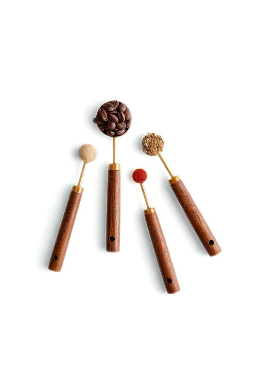 Four measuring spoons with wooden handles and brass-toned scoops, styled with dry ingredients