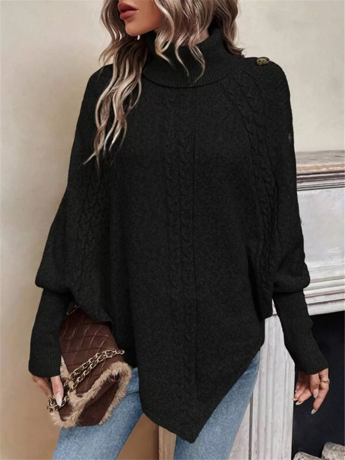 Black cable-knit turtleneck cape sweater. Front view with handbag, model in blue jeans. Styling accessories are not included.