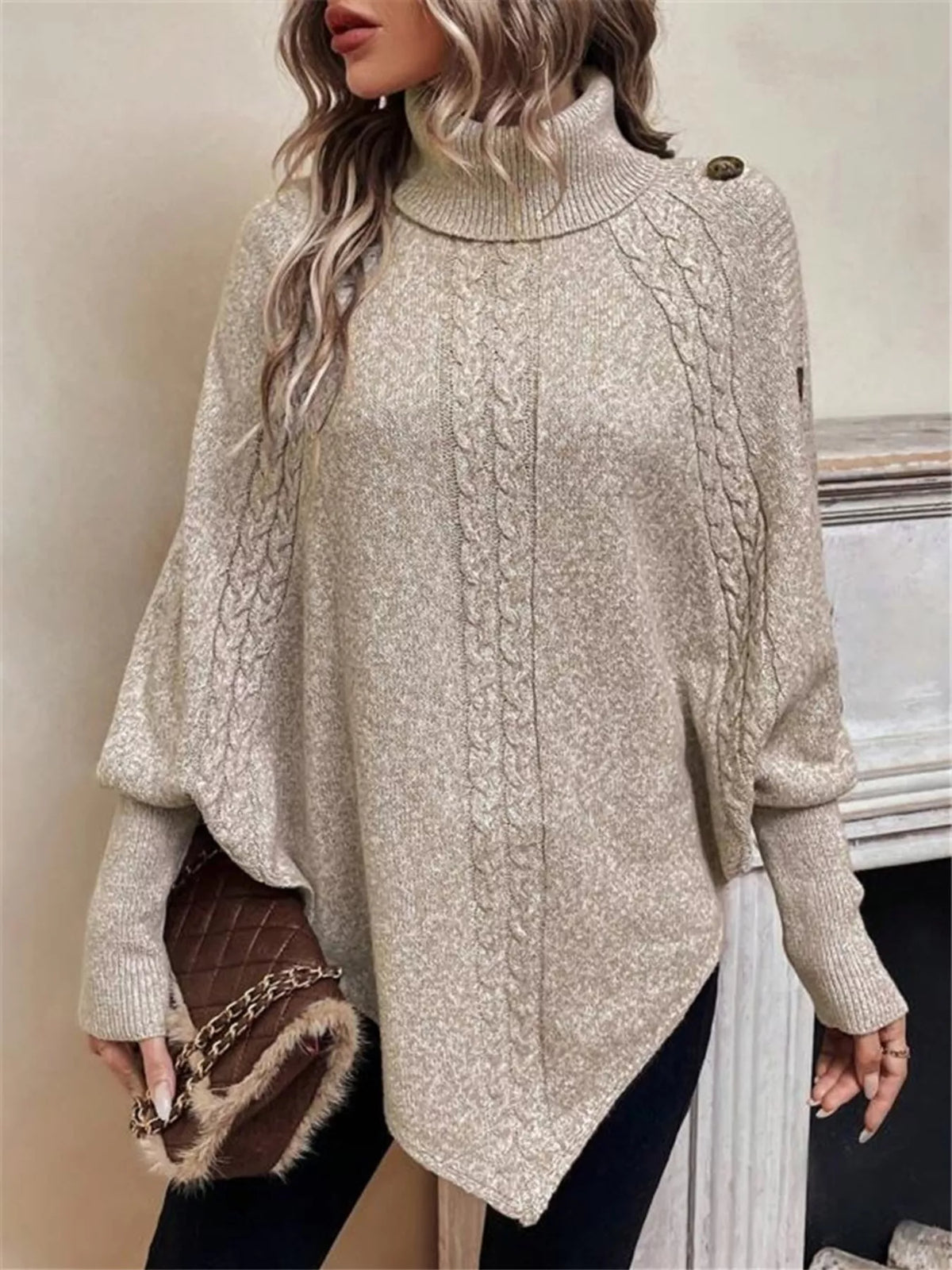 Warm beige/oatmeal cable-knit turtleneck cape sweater. Front three-quarter view, model in black pants. Styling accessories are not included.