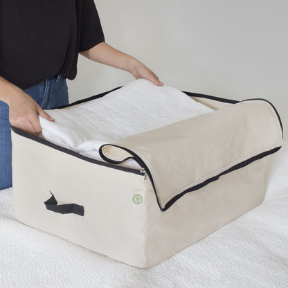 A natural canvas comforter storage bag open while a person places bedding inside