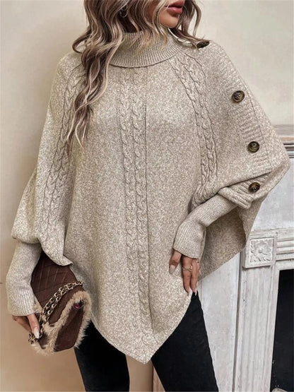 Warm beige/oatmeal cable-knit turtleneck cape sweater. Front/side view emphasizing cape drape and sleeve buttons. Styling accessories are not included.