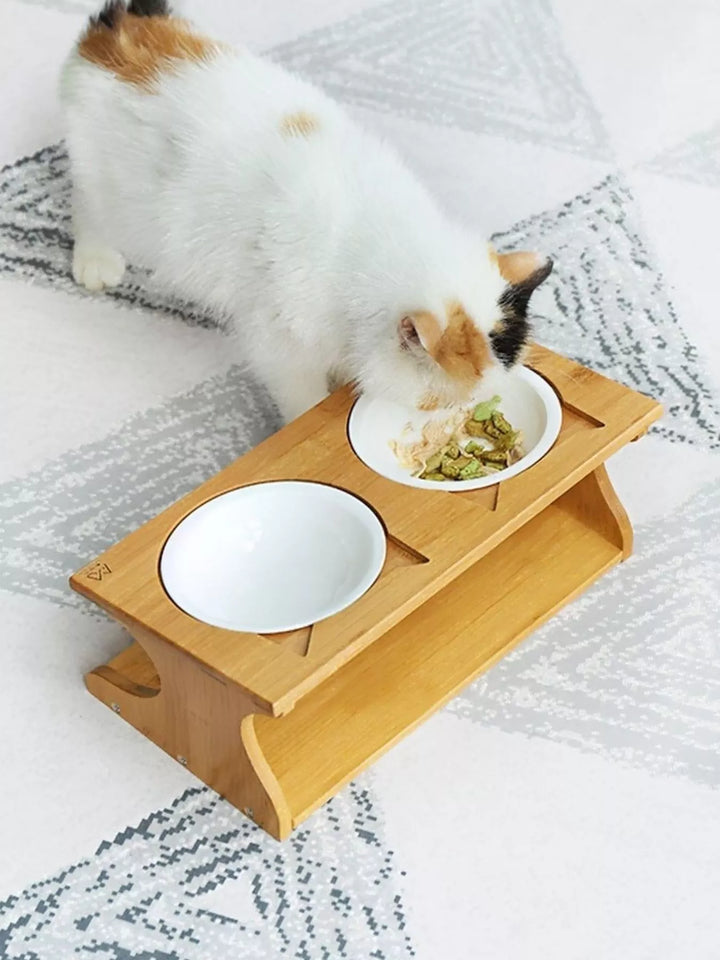 Elevated double pet-bowl set with two white ceramic bowls in a wooden stand, shown with a cat and food for illustration.