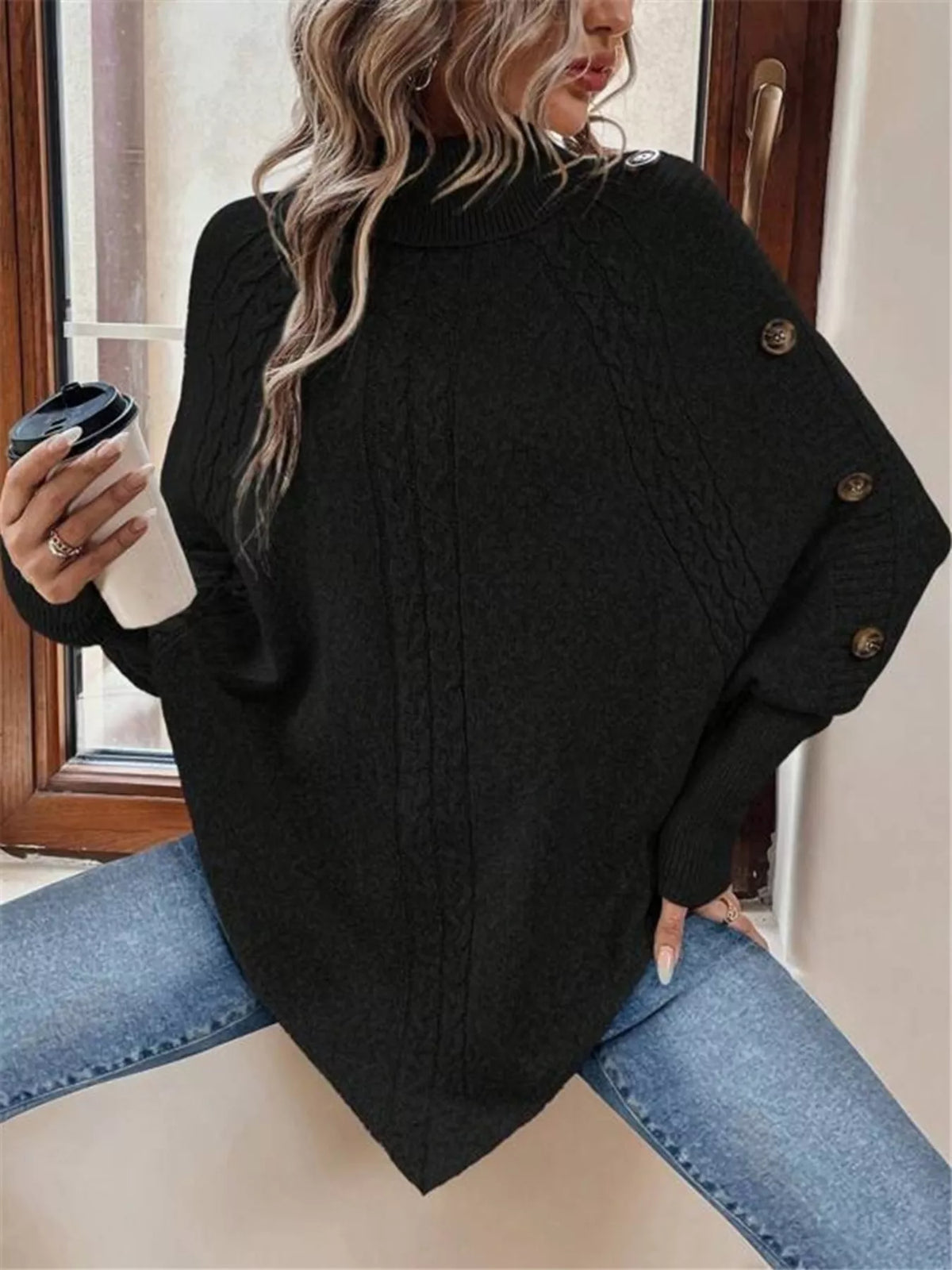 Black cable-knit turtleneck cape sweater. Seated front/side view, model in blue jeans. Styling accessories are not included.