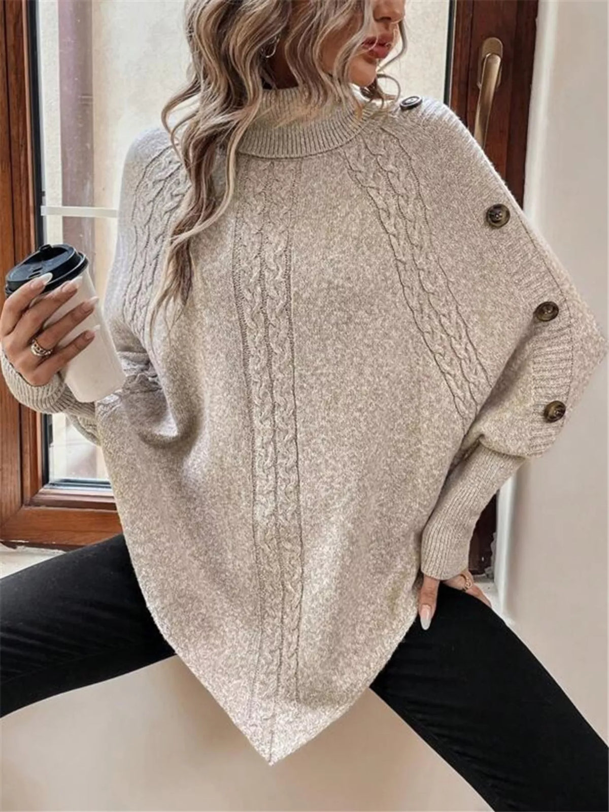 Warm beige/oatmeal cable-knit turtleneck cape sweater. Seated pose showing cape drape, model in black pants. Styling accessories are not included.
