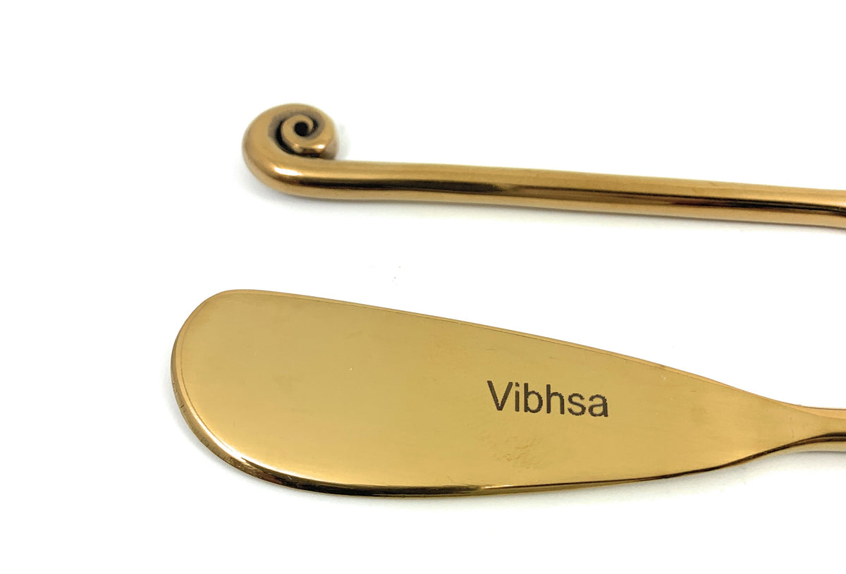Close-up of a Vibhsa gold-finish butter spreader blade and curled handle; this listing includes six matching spreaders.