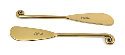 Two views of Vibhsa gold-finish butter spreaders with curled handles on a white background; sold as a set of six.