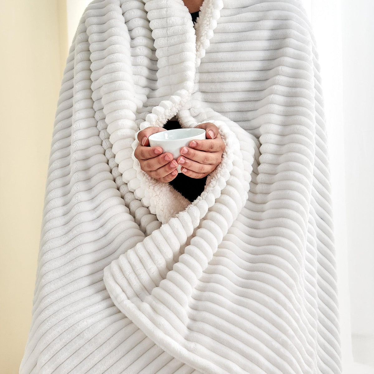 Person wrapped in the white ribbed sherpa blanket while holding a mug; one blanket only.