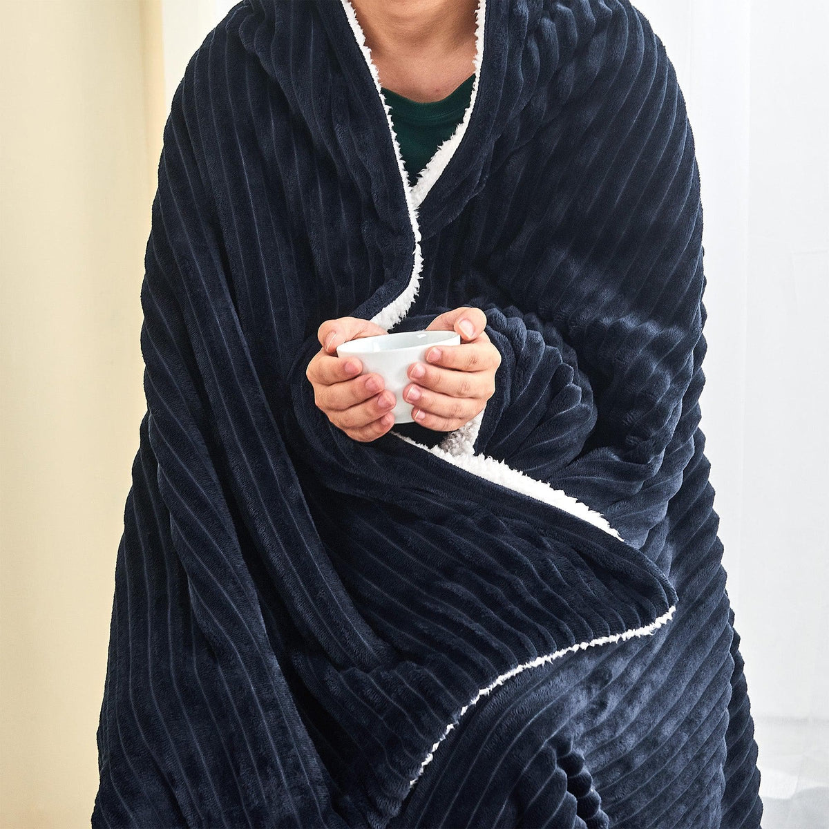 Person wrapped in the navy ribbed sherpa blanket while holding a mug; one blanket only.