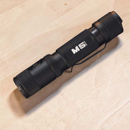 Black Powertac M5 Gen3 flashlight with pocket clip on a light wood surface